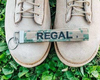 Puede incluir: Un par de botas de cuero color canela con cordones y un llavero con estampado de camuflaje con la palabra "REGAL" impresa.