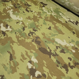 Peut inclure: Un rouleau de tissu présentant un motif camouflage multicolore. Le design se compose de formes irrégulières et superposées dans des tons de vert olive, beige clair, brun foncé et blanc cassé, créant un aspect texturé et tacheté sur la surface.