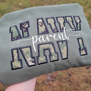 Op de afbeelding: Een olijfgroen sweatshirt met de geborduurde tekst "AF AITN parent" in een digitaal camouflagepatroon met zwarte omlijningen. Het woord "parent" is geschreven in een wit cursief lettertype tussen de twee regels met blokletters.
