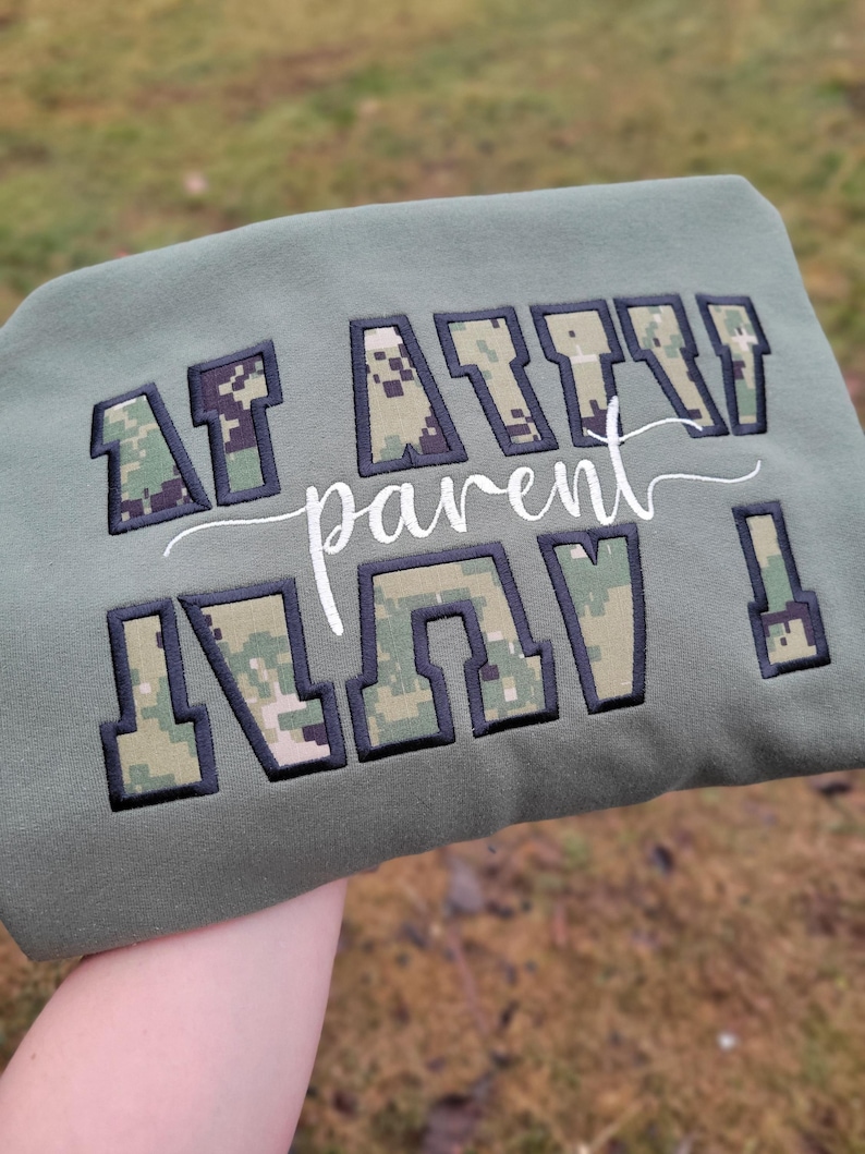 Op de afbeelding: Een olijfgroen sweatshirt met de geborduurde tekst "AF AITN parent" in een digitaal camouflagepatroon met zwarte omlijningen. Het woord "parent" is geschreven in een wit cursief lettertype tussen de twee regels met blokletters.