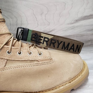 Peut inclure: Une botte en cuir beige avec une boucle noire et une sangle camouflage avec le texte "BERRYMAN" en noir.