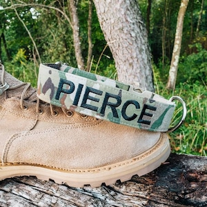 May include: A camouflage dog collar with black stitching and the name "PIERCE" embroidered in black.
