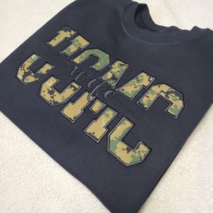 Op de afbeelding: Een donkergrijs sweatshirt met ronde hals en grote blokletters met een digitaal camouflagepatroon in groen, beige en bruin. De tekst "USMC" is in blokletters weergegeven met het woord "wife" in een cursief lettertype in het midden.