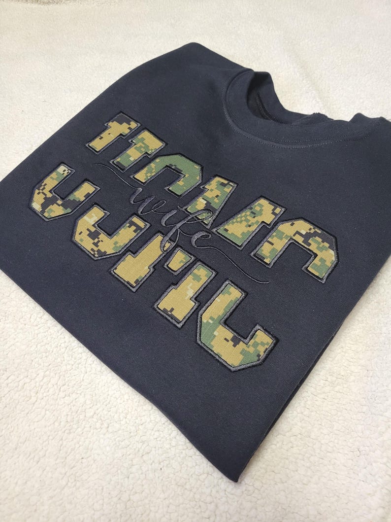Op de afbeelding: Een donkergrijs sweatshirt met ronde hals en grote blokletters met een digitaal camouflagepatroon in groen, beige en bruin. De tekst "USMC" is in blokletters weergegeven met het woord "wife" in een cursief lettertype in het midden.