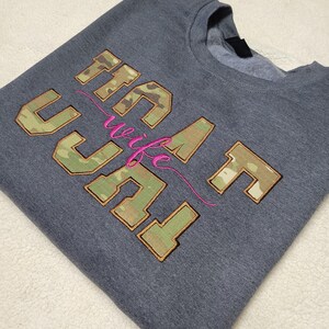 Op de afbeelding: Een antracietgrijze sweater met ronde hals en het geborduurde opschrift "NAVY wife" in blokletters. De letters zijn gevuld met een groen, bruin en beige camouflagepatroon en bruin omlijnd. Het woord "wife" is in cursieve felroze draad.