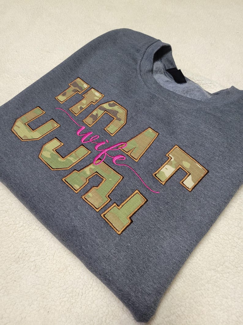 Op de afbeelding: Een antracietgrijze sweater met ronde hals en het geborduurde opschrift "NAVY wife" in blokletters. De letters zijn gevuld met een groen, bruin en beige camouflagepatroon en bruin omlijnd. Het woord "wife" is in cursieve felroze draad.