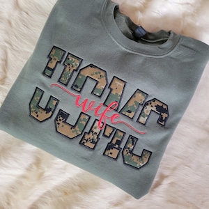 Op de afbeelding: Een olijfgroene sweater met ronde hals en de tekst "MARIN WIFE" in blokletters met een digitale camouflagevulling. Het woord "wife" is in een rood cursief lettertype over het midden van de blokletters geborduurd.