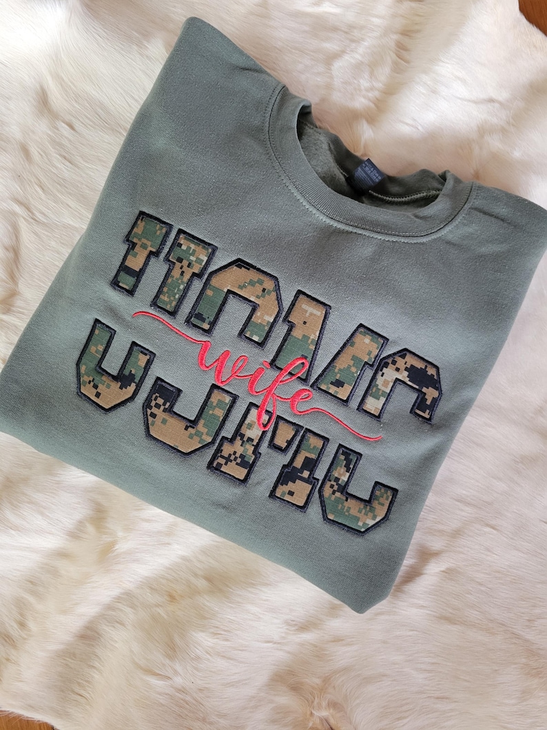 Op de afbeelding: Een olijfgroene sweater met ronde hals en de tekst "MARIN WIFE" in blokletters met een digitale camouflagevulling. Het woord "wife" is in een rood cursief lettertype over het midden van de blokletters geborduurd.