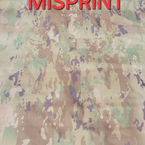 May include: A fabric featuring a camouflage pattern in shades of tan, light green, and muted purple. The word MISPRINT is printed in bold red capital letters across the center of the fabric, which has a visible woven texture throughout the surface.