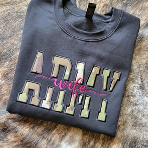 Op de afbeelding: Een zwart sweatshirt met ronde hals en de tekst ARMY WIFE geborduurd op de voorzijde. Het woord ARMY staat in blokletters met een camouflagepatroon, terwijl het woord wife in een roze cursief lettertype tussen de regels is geplaatst.