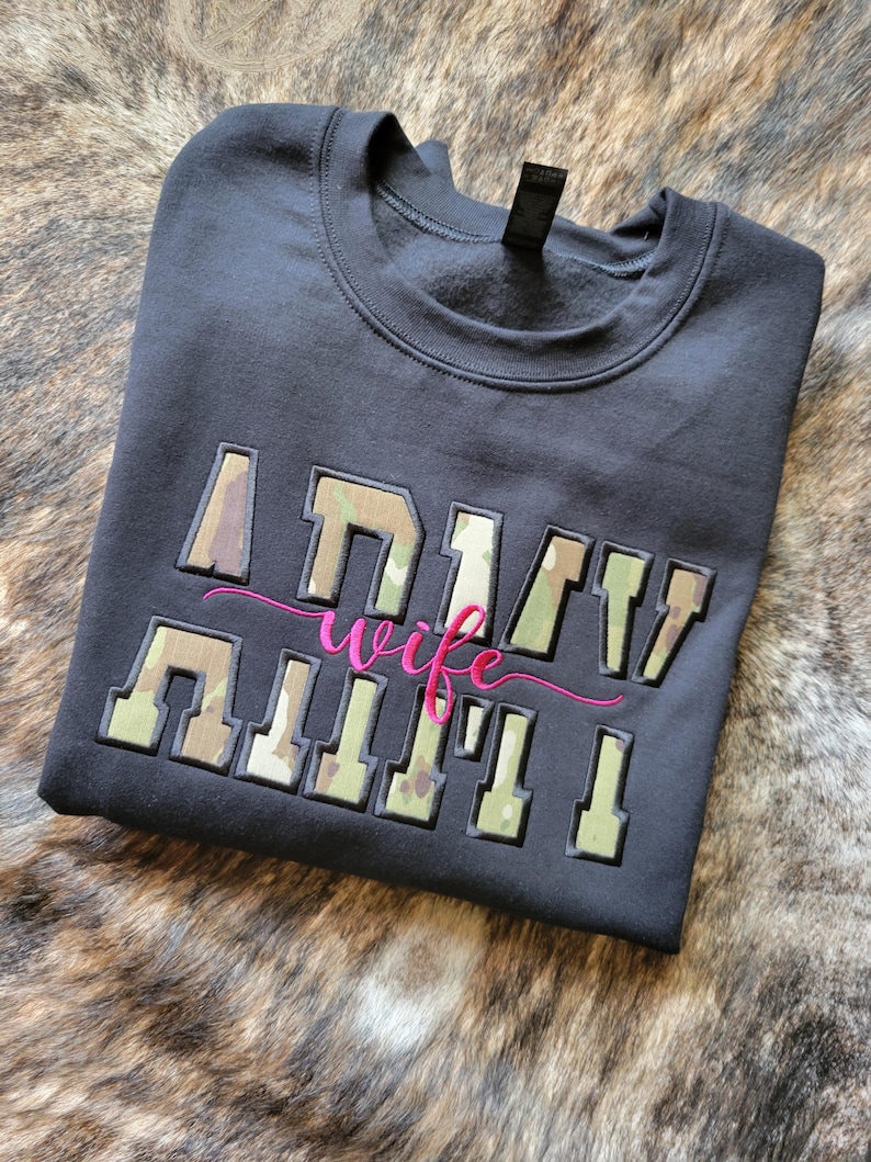 Op de afbeelding: Een zwart sweatshirt met ronde hals en de tekst ARMY WIFE geborduurd op de voorzijde. Het woord ARMY staat in blokletters met een camouflagepatroon, terwijl het woord wife in een roze cursief lettertype tussen de regels is geplaatst.