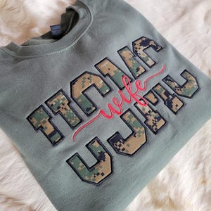 Op de afbeelding: Een olijfgroene sweater met ronde hals en grote blokletters met een digitaal camouflagepatroon. Het woord "wife" is in rode cursieve letters geborduurd over het midden van de blokletters die het woord "MARINE" vormen.