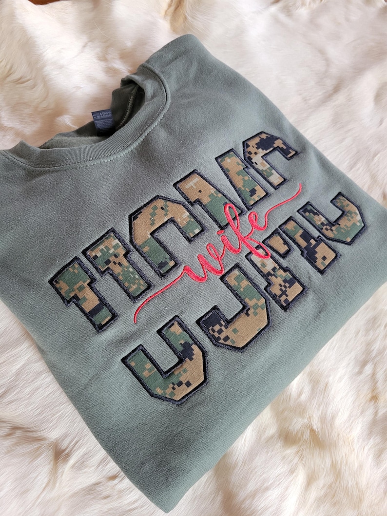 Op de afbeelding: Een olijfgroene sweater met ronde hals en grote blokletters met een digitaal camouflagepatroon. Het woord "wife" is in rode cursieve letters geborduurd over het midden van de blokletters die het woord "MARINE" vormen.
