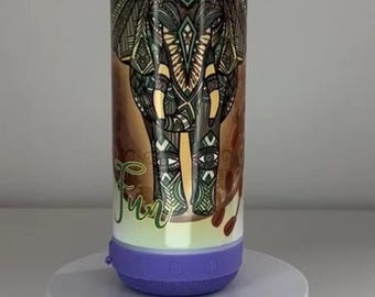 May include: A cylindrical wireless speaker featuring a detailed elephant illustration with intricate patterns in shades of brown, green, and black. The name Orla and the word Fun are printed in green script. The base is purple with control buttons.