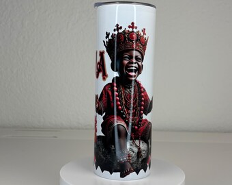 May include: A white cylindrical tumbler featuring a graphic print of a laughing child wearing a red and gold crown and red traditional attire with beaded necklaces. The child is seated on a dark stone-like base against a plain white background.