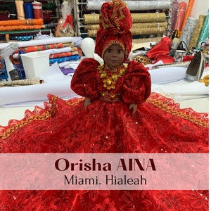 May include: A red and gold doll dressed in a traditional Orisha Aina costume. The doll is wearing a red dress with gold trim and a red head wrap with gold accents. The doll is sitting on a table with a white tablecloth. The text "Orisha AINA" and "Miami. Hialeah" is visible in the image.