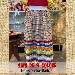 May include: A long, colorful skirt with a plaid pattern in yellow, red, and blue. The skirt features horizontal bands of vibrant ribbons in various colors, including red, blue, orange, and purple. The text "SAYA DE 9 COLORE Trajes Orishas Barbara" is displayed at the bottom.