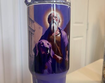 May include: A purple stainless steel tumbler featuring a graphic of a bearded man with a halo and a dog. The tumbler has a clear lid with a purple straw and the text San Lázaro printed in black script near the base. The design includes architectural elements.