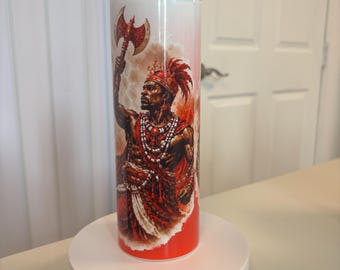 May include: A cylindrical tumbler featuring a vibrant graphic print of a warrior figure adorned in traditional red and white attire, wearing a feathered headdress and beaded necklaces, while holding a large battle axe raised high in the air.