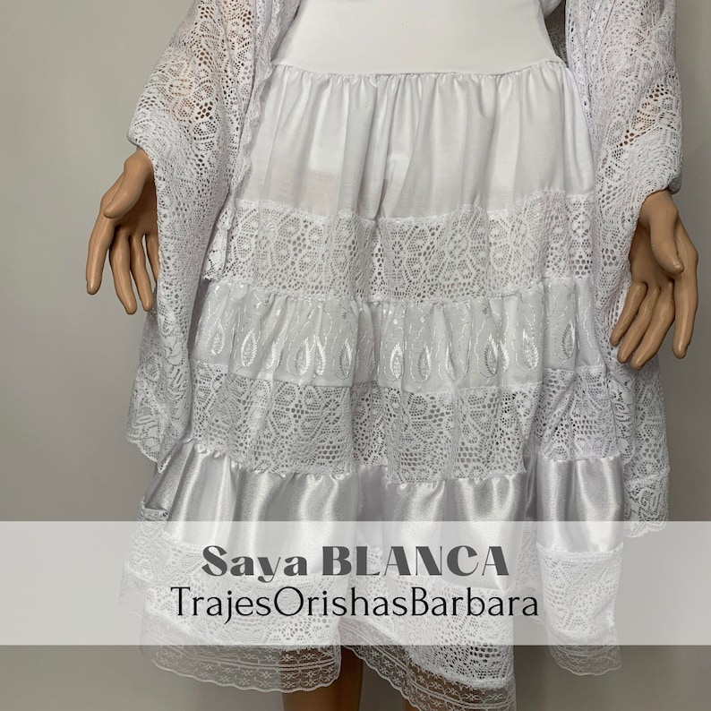 May include: White lace skirt with a satin trim and embroidered details. The skirt is layered and has a flowing silhouette. The text "Saya BLANCA" and "TrajesOrishasBarbara" is printed on the bottom of the skirt.
