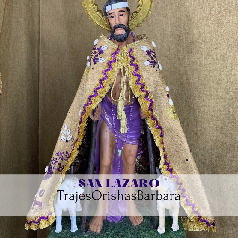 Puede incluir: Estatua de San L&aacute;zaro, figura religiosa, con capa beige con ribetes morados y dorados, y vestimenta morada. La estatua tiene una cinta blanca en la cabeza y un halo dorado. Dos perros blancos est&aacute;n en la base. El texto "SAN LAZARO TrajesOrishasBarbara" est&aacute; en la parte inferior.