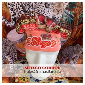 May include: A red, gold, and black patterned chef's hat with a crown and the word "Shango" in gold lettering. The hat is on a white mannequin head against a background of leopard print fabric.