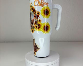 May include: A white stainless steel tumbler with a handle and a clear lid, featuring a printed design of yellow sunflowers, butterflies, and a djembe drum. The name Oshun is written in orange cursive script near the top of the drinkware.