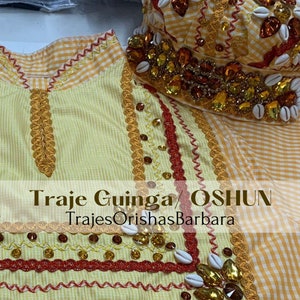 May include: A yellow and white gingham fabric with gold and red trim. The fabric is decorated with gold beads and white seashells. The text "Traje Guinga/ OSHUN TrajesOrishasBarbara" is printed on the fabric.