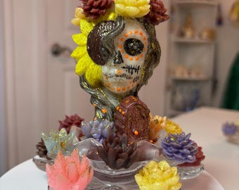 May include: A decorative candle shaped like a skull with a floral crown, featuring yellow, pink, and dark red wax succulents at the base. The skull has black and orange painted details, glitter accents, and a small brown coffin figure in front.