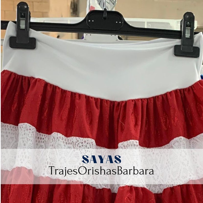 May include: Red skirt with white lace trim and a white waistband. The skirt has the text "SAYAS TrajesOrishasBarbara" printed on the bottom.