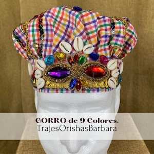 May include: A colorful checkered hat with a gold trim and a band of white seashells and colorful jewels. The hat is labeled "GORRO de 9 Colores. TrajesOrishasBarbara".