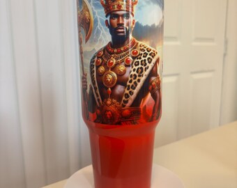 May include: A cylindrical tumbler with a red base and a clear lid featuring a red straw. The upper portion displays a graphic of a king wearing a crown, a leopard print cape, and gold jewelry, holding a battle axe against a stormy sky background.