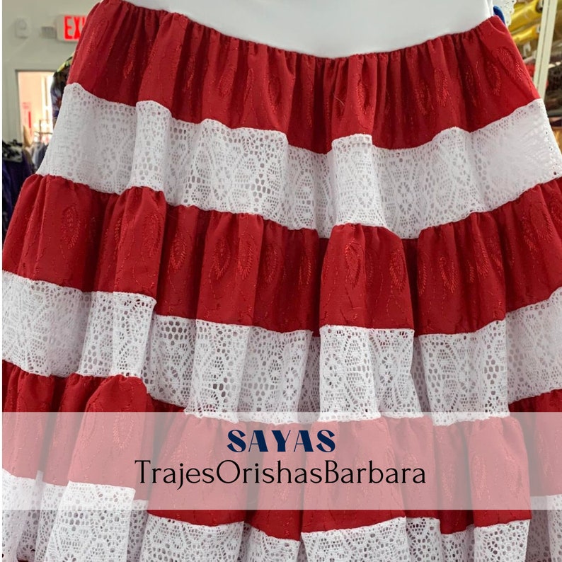 May include: Red and white layered skirt with white lace trim. The skirt has the text "SAYAS TrajesOrishasBarbara" printed on the bottom.