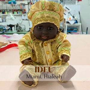 May include: A small doll wearing a gold and green outfit with a gold crown. The doll has a black face and is sitting on a pink surface. The text "IDEU Miami. Hialeah" is printed below the doll.