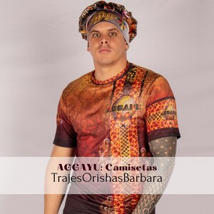 May include: A man wearing a colorful patterned shirt with the text "AGGAYU: Camisetsa TrajesOrishasBarbara" on the bottom. He is also wearing a white satin head wrap with the word "Mayara" written on it.