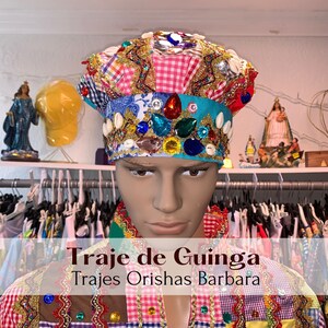 May include: A colorful, multi-patterned hat with a crown of jewels and shells. The hat is worn by a mannequin with a colorful, multi-patterned shirt. The text "Traje de Guinga" and "Trajes Orisha Barbara" is visible on the image.