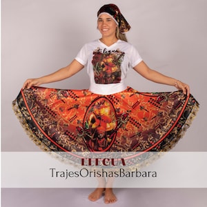 Puede incluir: Una mujer con una camisa blanca con el texto "Eleggua" impreso, una falda colorida con ribete negro y dorado, y una pañoleta de cabeza blanca y negra. La falda tiene un gran diseño ovalado con una figura en el centro. El texto "Eleggua TrajesOrichasBarbara" está impreso en la parte inferior de la imagen.