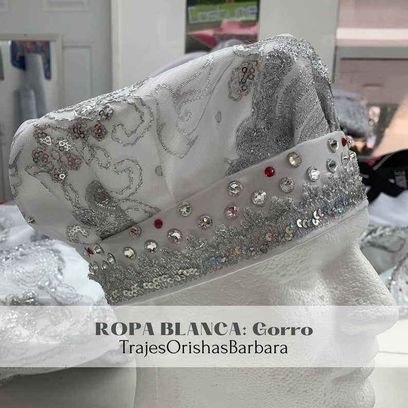 May include: White hat with silver and red embellishments. The hat is decorated with sequins and rhinestones. The text "ROPA BLANCA: Gorro TrajesOrishasBarbara" is printed on the hat.
