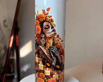 May include: A cylindrical white tumbler featuring a colorful graphic print of a person with sugar skull face paint, wearing orange marigold flowers in their hair, surrounded by lit candles and autumn leaves, with a clear plastic lid on top.