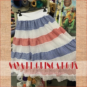 May include: A white, blue, and red gingham skirt with a white lace trim. The skirt has a white elastic waistband and is layered with white lace. The skirt is labeled "Saya de Ginga Roja Trajes Orishas Barbara".
