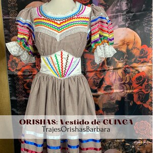 May include: A brown and white gingham dress with colorful ribbon trim and embroidery. The dress has a white lace trim on the sleeves and a white waistband with colorful embroidery. The dress is labeled "ORISHAS: Vestido de GUINGA TrajesOrishasBarbara".