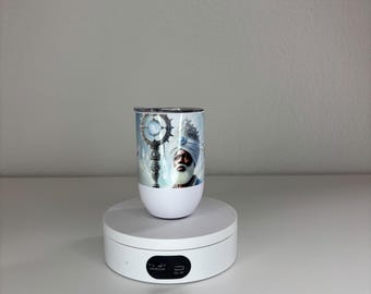 May include: A white tumbler featuring a graphic print of a person wearing a turban and ornate headpiece, holding a staff with a glowing orb. The tumbler has a metallic rim and lid, and is displayed on a white circular rotating stand.