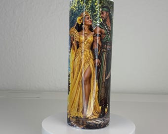 May include: A cylindrical tumbler featuring a full-wrap graphic print of a person in a flowing yellow gown with a fan and a person in green warrior attire standing together in a forest setting. The design includes intricate gold jewelry and details.