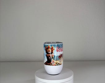 May include: A white cylindrical tumbler featuring a graphic print of a person wearing a gold crown and leopard print clothing. The design includes red text and crown imagery against a blue background. The tumbler sits on a white rotating display stand.