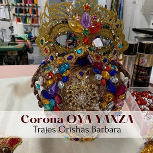May include: A gold crown with a jeweled mask on top. The crown is decorated with colorful jewels and rhinestones. The text "Corona OYA YANZA Trajes Orishas Barbara" is visible at the bottom of the image.