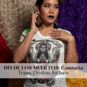 May include: A white t-shirt featuring a black and white illustration of Santa Muerte, a female skeletal figure adorned with a crown of roses. The text "Santa Muerte" is printed above the illustration. The t-shirt is being worn by a person with dark hair and makeup. The background is a purple and gold fabric.