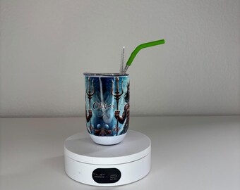 May include: A cylindrical tumbler featuring a graphic print of a figure holding a trident with the text Olokun. The tumbler includes a clear lid and a bright green bent straw. The design displays blue and dark tones with aquatic imagery.