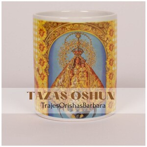 May include: White ceramic mug with a colorful image of a religious figure in a yellow and blue robe, surrounded by sunflowers. The text "TAZAS OSHUN" and "TrajesOrishasBarbara" are printed on the mug.