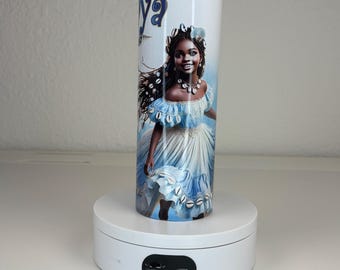May include: A cylindrical tumbler featuring a graphic illustration of a person with dark skin and long dark hair, wearing a light blue dress adorned with white cowrie shells. The text "ya" is visible at the top. The tumbler has a metallic rim.