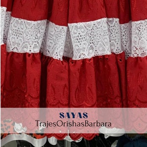 May include: Red skirt with white lace trim. The skirt has a scalloped edge and is embroidered with a floral design. The text "SAYAS TrajesOrishasBarbara" is visible at the bottom of the image.